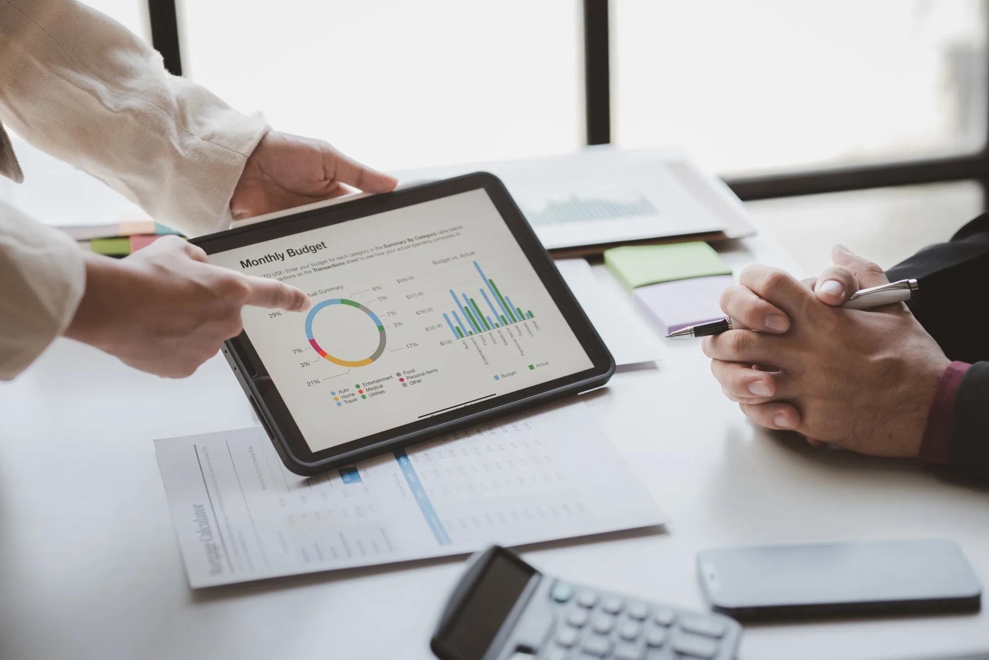 Two people in a business setting discussing financial data. One person is pointing at a tablet displaying a monthly budget chart with a pie chart and bar graph. The other person is holding a pen and taking notes. Documents, sticky notes, and office supplies are on the table.