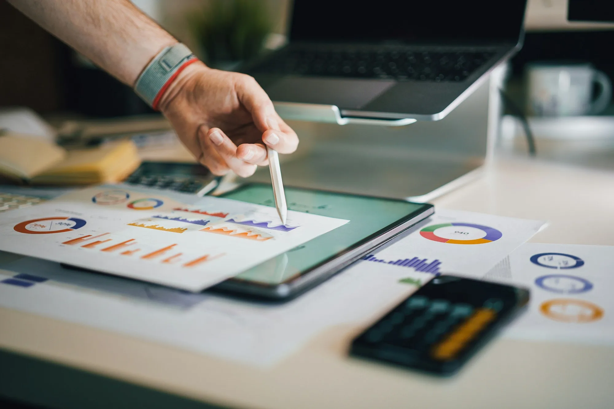 Hand using stylus on tablet displaying colorful charts and graphs, with a laptop and printed charts on desk.