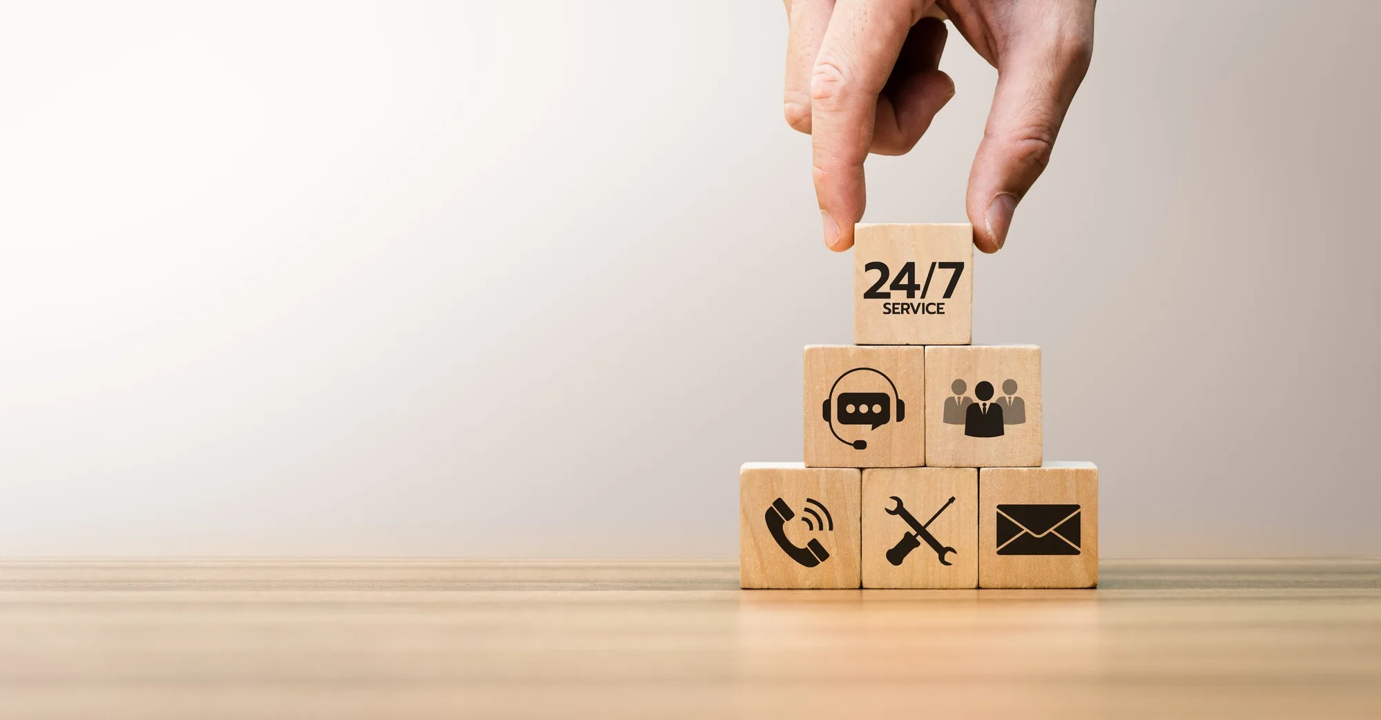 A hand stacking wooden blocks with icons and text representing 24/7 service, customer support, communication, tools, and email, arranged in a pyramid shape on a wooden surface.