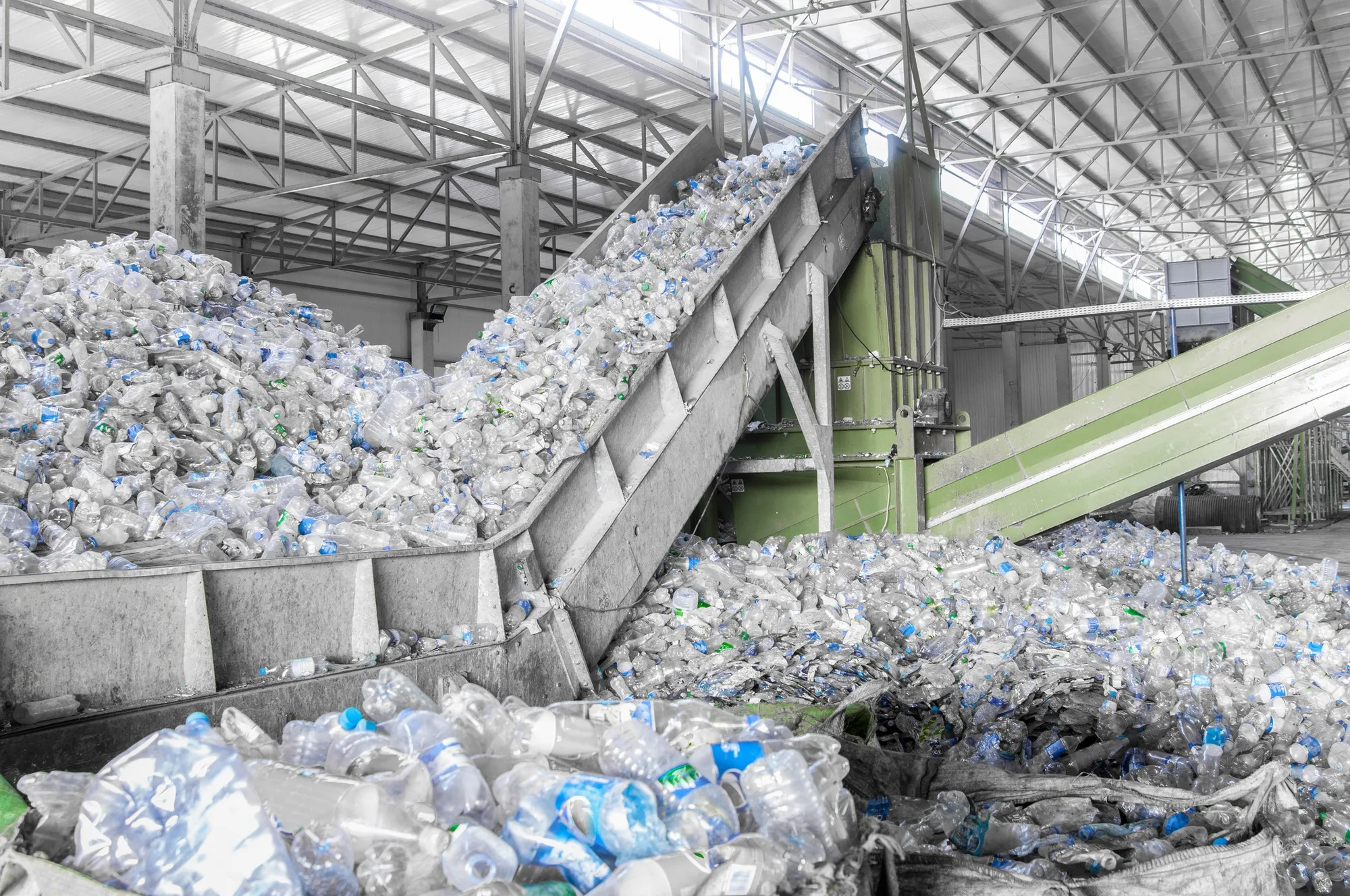 A large conveyor belt in a recycling facility is sorting plastic bottles, with many crushed and uncrushed bottles on it, under a metal roof structure.