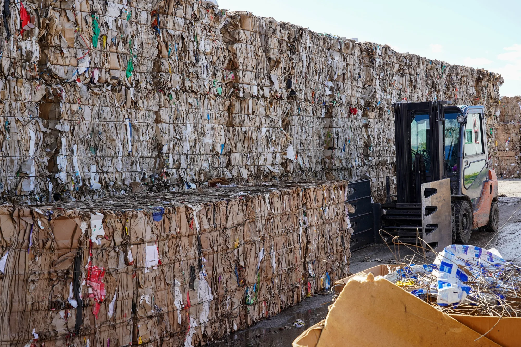 Stacks of compressed paper or cardboard at a recycling center with a forklift nearby, under a partly cloudy sky.